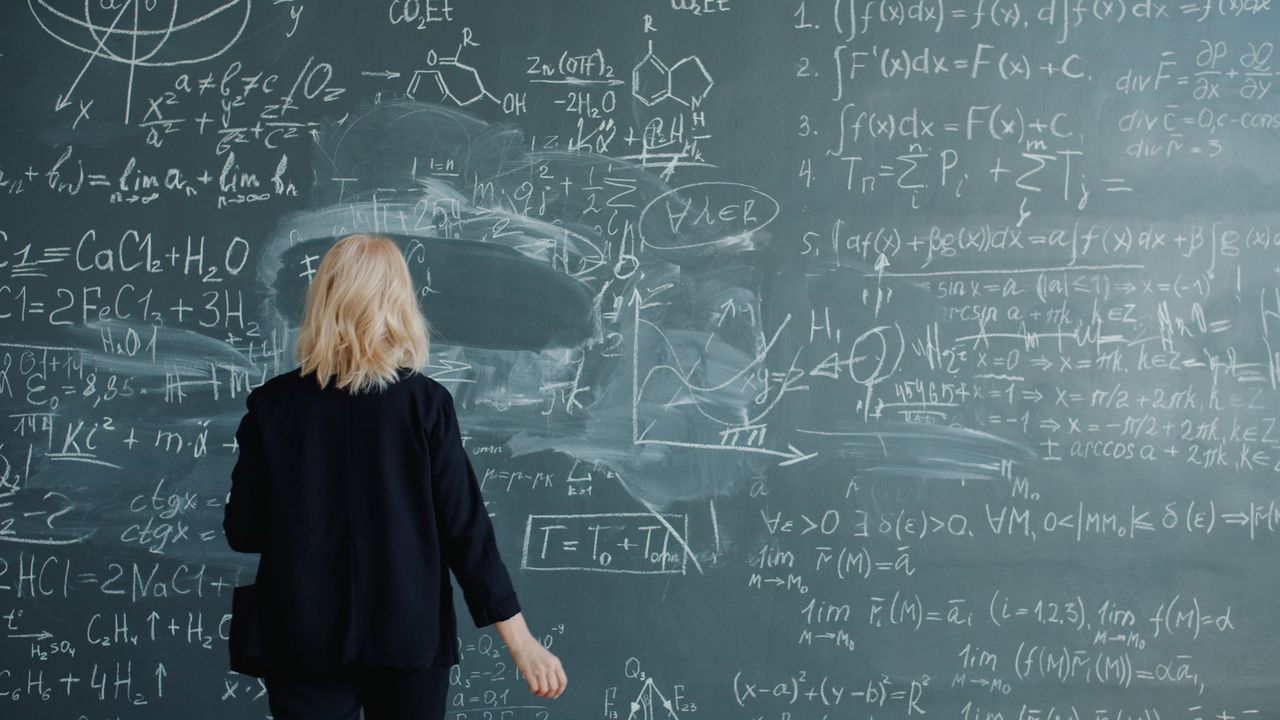 about-us Woman writing on chalkboard filled with complex mathematical equations.